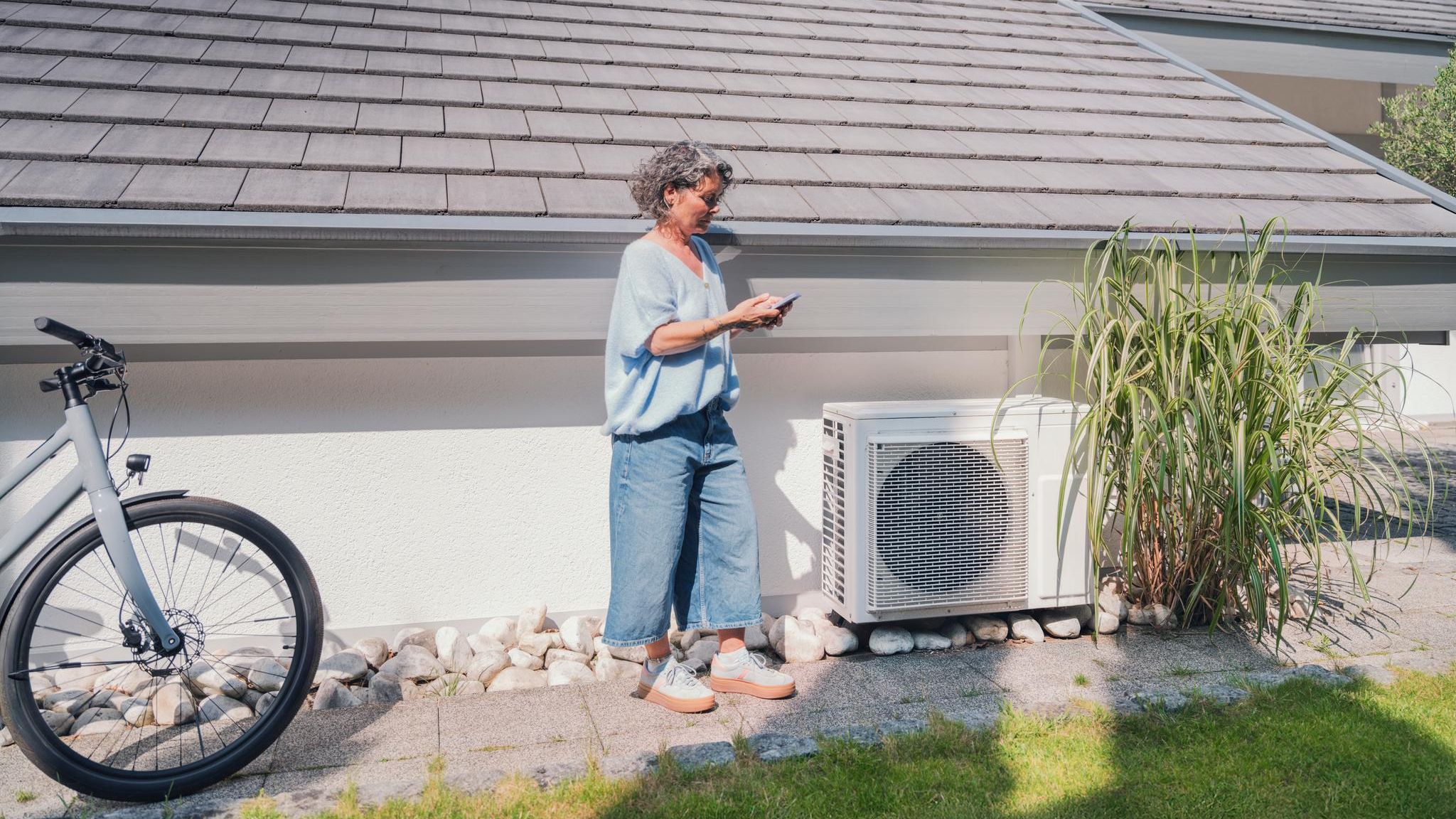 Wärmepumpenstrom-Vergleich: Eine Frau checkt ihr Handy neben ihrer Wärmepumpe