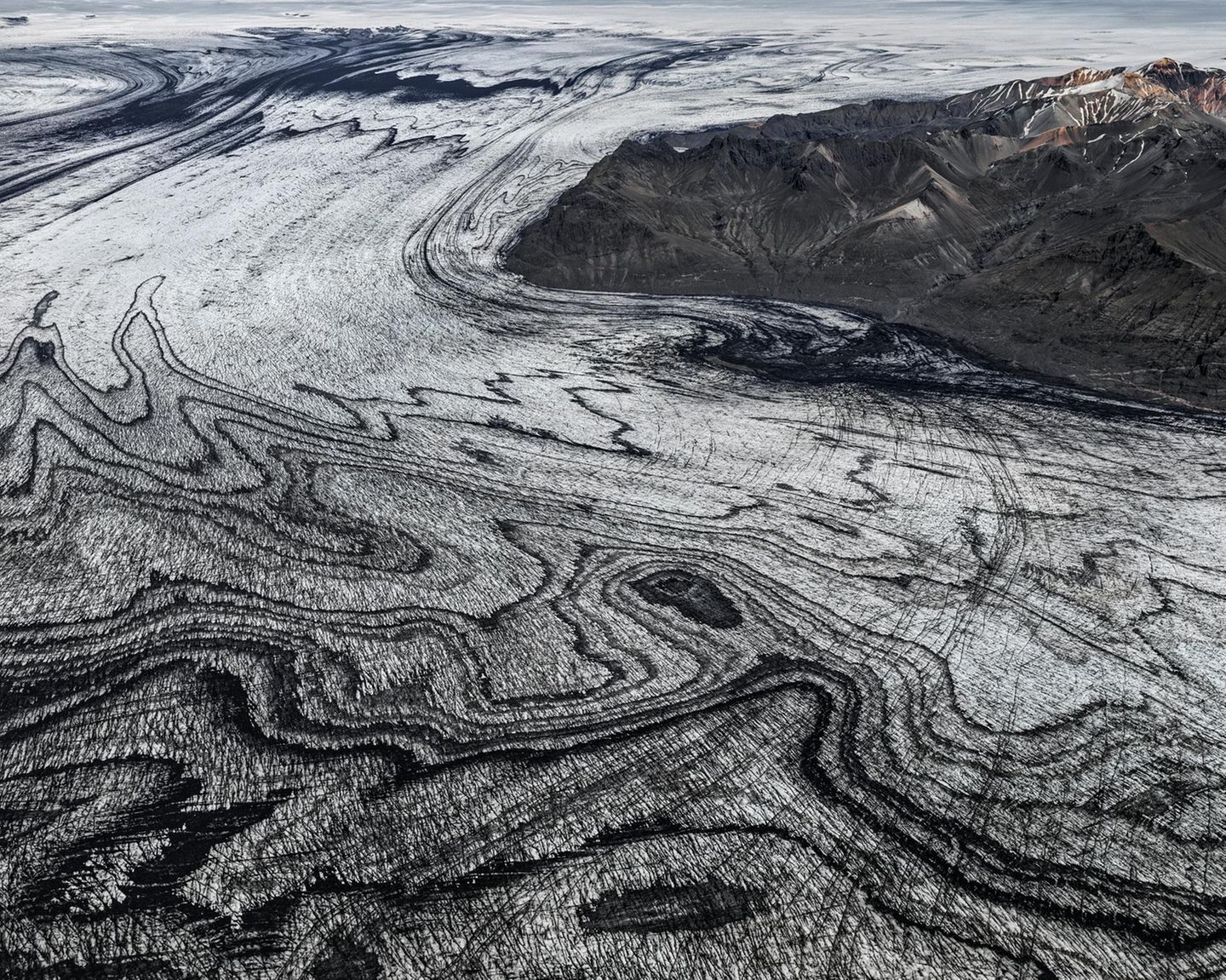 Wenn sich Gletscherströme ihren Weg durch das Gebirge bahnen, verschlägt ihre schiere Kraft den Betrachtenden meist den Atem. Die schwarze Vulkanasche, die das Eis des Skeidararjökull hier marmoriert, zeugt jedoch von einem noch gewaltigeren Naturspektakel: dem Wechselspiel zwischen Schnee und Feuer, Sonne und Wind. "Die Vulkaneruptionen liefern die schwarze Asche, welche die Gletscher überdeckt. Darauf fällt frischer Schnee, der allmählich zu Eis komprimiert wird. So legt sich weiße auf schwarze Schicht, viele Male", beschreibt die Geologin Angelika Jung-Hüttl das Phänomen. Je weiter sich der Gletscher seinen Weg ins Tal bahnt, desto verwobener sind die beiden Schichten, desto schwungvoller die Muster