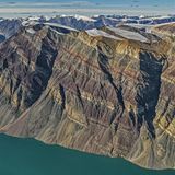 Wie bei einer Schichttorte haben sich die unterschiedlich gefärbten Gesteinsschichten im Laufe der Jahrmillionen zu dem Gebirgsstock gestapelt, der heute am Rande des Kong-Oscar-Fjords in Ostgrönland thront. Der schroffe Fels überragt den Fjord um 1900 Meter und zeugt noch heute von seiner einstigen Lage am Rande eines urzeitlichen Ozeans. Auf dessen Grund lagerten sich in verschiedenen geologischen Zeiten verschiedenfarbige Materialien ab. Dass diese Schichtung noch heute zu bewundern ist, ist der Tatsache zu verdanken, dass das Gebirge während der Kaledonischen Gebirgsbildung vor 400 Millionen Jahren – im Gegensatz zu anderen Bergzügen – nicht gefaltet, sondern lediglich angehoben, leicht gekippt und in Blöcke zerlegt wurde