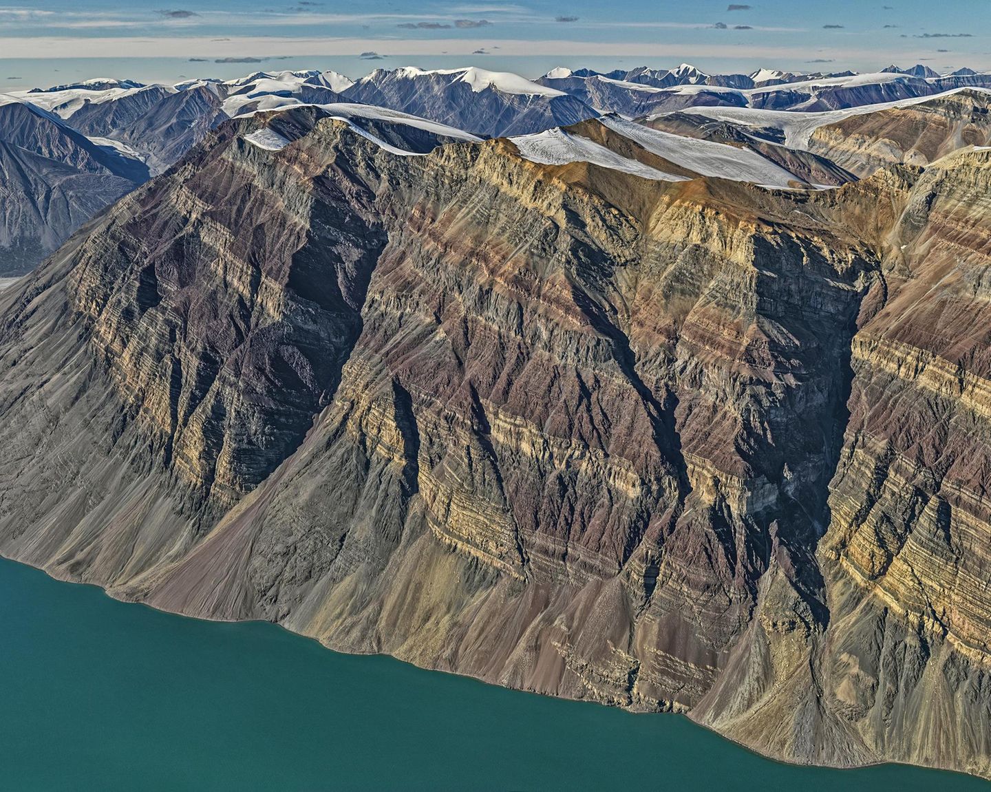 Wie bei einer Schichttorte haben sich die unterschiedlich gefärbten Gesteinsschichten im Laufe der Jahrmillionen zu dem Gebirgsstock gestapelt, der heute am Rande des Kong-Oscar-Fjords in Ostgrönland thront. Der schroffe Fels überragt den Fjord um 1900 Meter und zeugt noch heute von seiner einstigen Lage am Rande eines urzeitlichen Ozeans. Auf dessen Grund lagerten sich in verschiedenen geologischen Zeiten verschiedenfarbige Materialien ab. Dass diese Schichtung noch heute zu bewundern ist, ist der Tatsache zu verdanken, dass das Gebirge während der Kaledonischen Gebirgsbildung vor 400 Millionen Jahren – im Gegensatz zu anderen Bergzügen – nicht gefaltet, sondern lediglich angehoben, leicht gekippt und in Blöcke zerlegt wurde