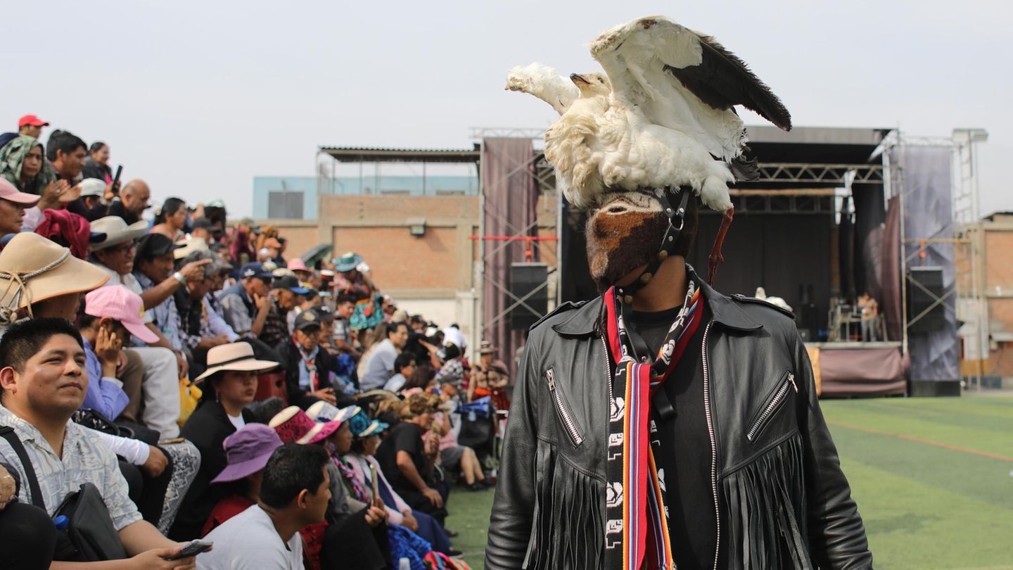 Ein Mensch mit einer Maske und einem Hut, bestehend aus einem ausgestopften Vogel, steht vor Publikum