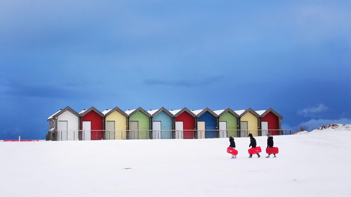 Blyth, Großbritannien. Auch im Nordosten Englands herrschen winterliche Bedingungen. Die bunten Hütten an diesem Strand nördlich von Newcastle upon Tyne sind geschlossen, drei junge Menschen sind aber mit Boards unterwegs. Diese werden sie aktuell wohl eher fürs Rodeln im Schnee als fürs Surfen im Wasser nutzen