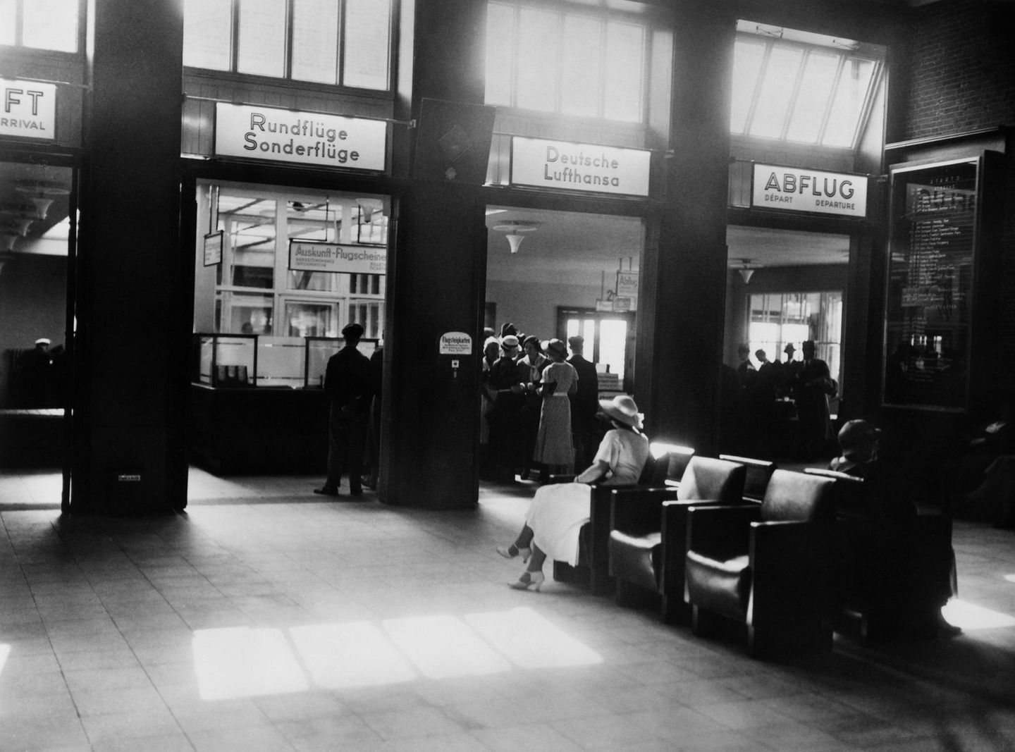Eine Abfertigungshalle der Lufthansa mit Sesseln und Menschen vor Schaltern, schwarz-weiß Foto