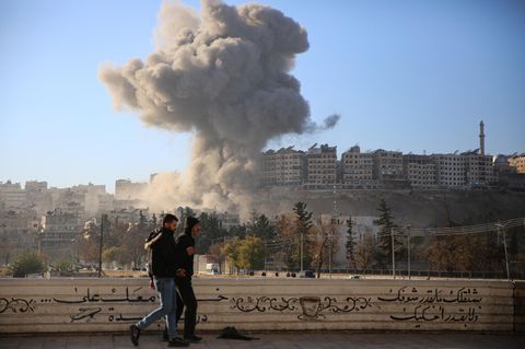 Zwei Männer laufen an einer Straße in Aleppo entlang, während weiter weg eine Rauchwolke aufsteigt