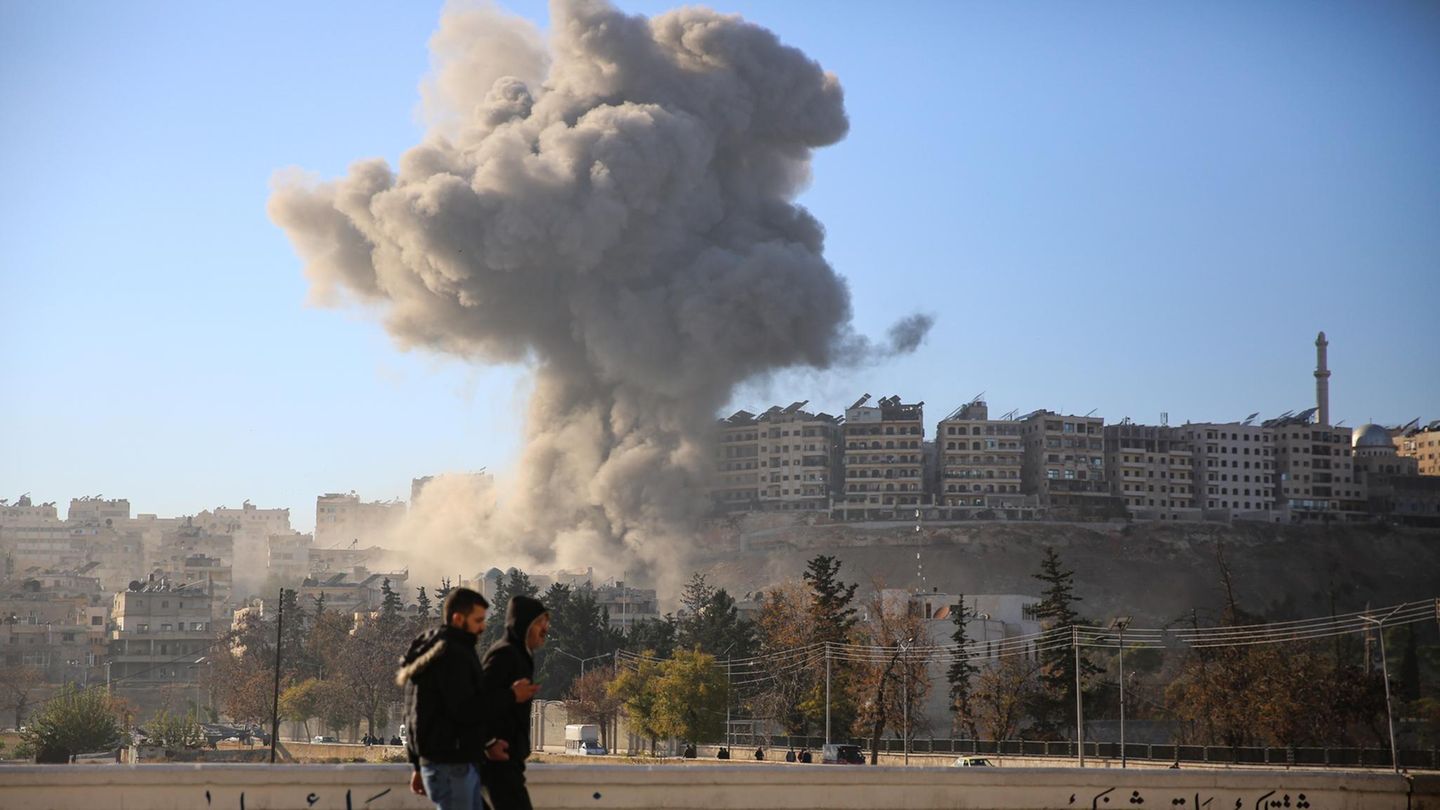 Zwei Männer laufen an einer Straße in Aleppo entlang, während weiter weg eine Rauchwolke aufsteigt