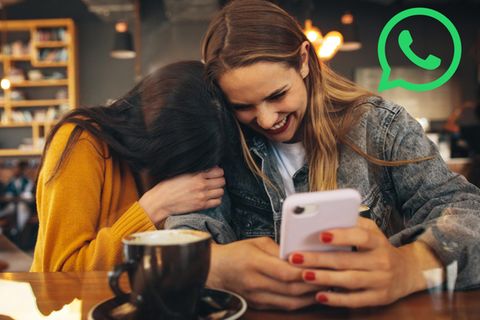 Zwei Frauen lachen über den Inhalt eines Smartphone-Bildschirms. Oben rechts ist das Whatsapp-Logo zu sehen
