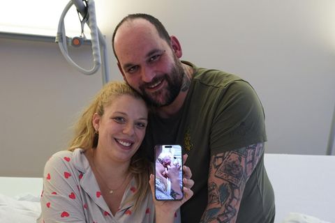 Eine Frau lächelt aus einem Krankenhaus-Bett in Hamburg, während sie ein Foto ihres Babys auf ihrem Smartphone zeigt