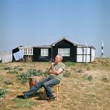 Eine Person sitzt entspannt auf einem Stuhl auf der Wiese, hinter ihr eine Holzhütte