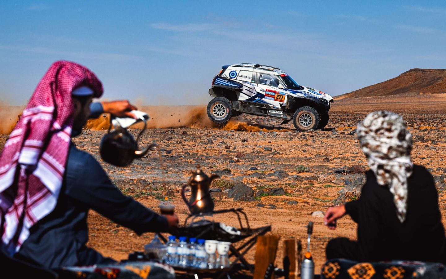 Zuschauer bereiten in Saudi-Arabien auf traditionelle Weise Tee zu, während im Hintergrund ein Rennwagen der Rallye Dakar Staub aufwirbelt. Der Wettbewerb hat eine lange Geschichte, hieß ursprünglich "Ralley Paris-Dakar", gastierte auch schon in Südamerika und wird inzwischen von Saudi-Arabien ausgerichtet. Wegen vieler Todesfälle, hoher Kosten und ökologischer Folgen steht die Veranstaltung seit Jahren in der Kritik