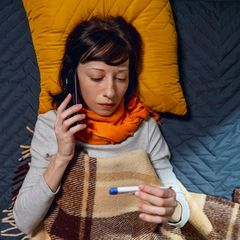 Eine Frau liegt auf dem Sofa, Schal um den Hals, Fieberthermometer in der Hand, und am Telefon für eine Krankschreibung