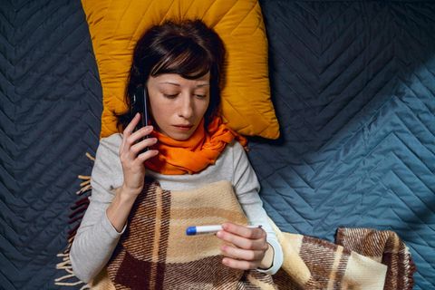 Eine Frau liegt auf dem Sofa, Schal um den Hals, Fieberthermometer in der Hand, und am Telefon für eine Krankschreibung