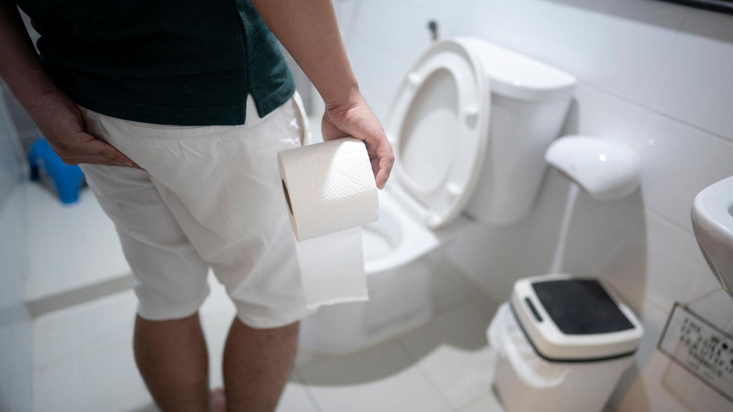 Ein Mann steht mit einer Rolle Klopapier in der Hand vor einer Toilette