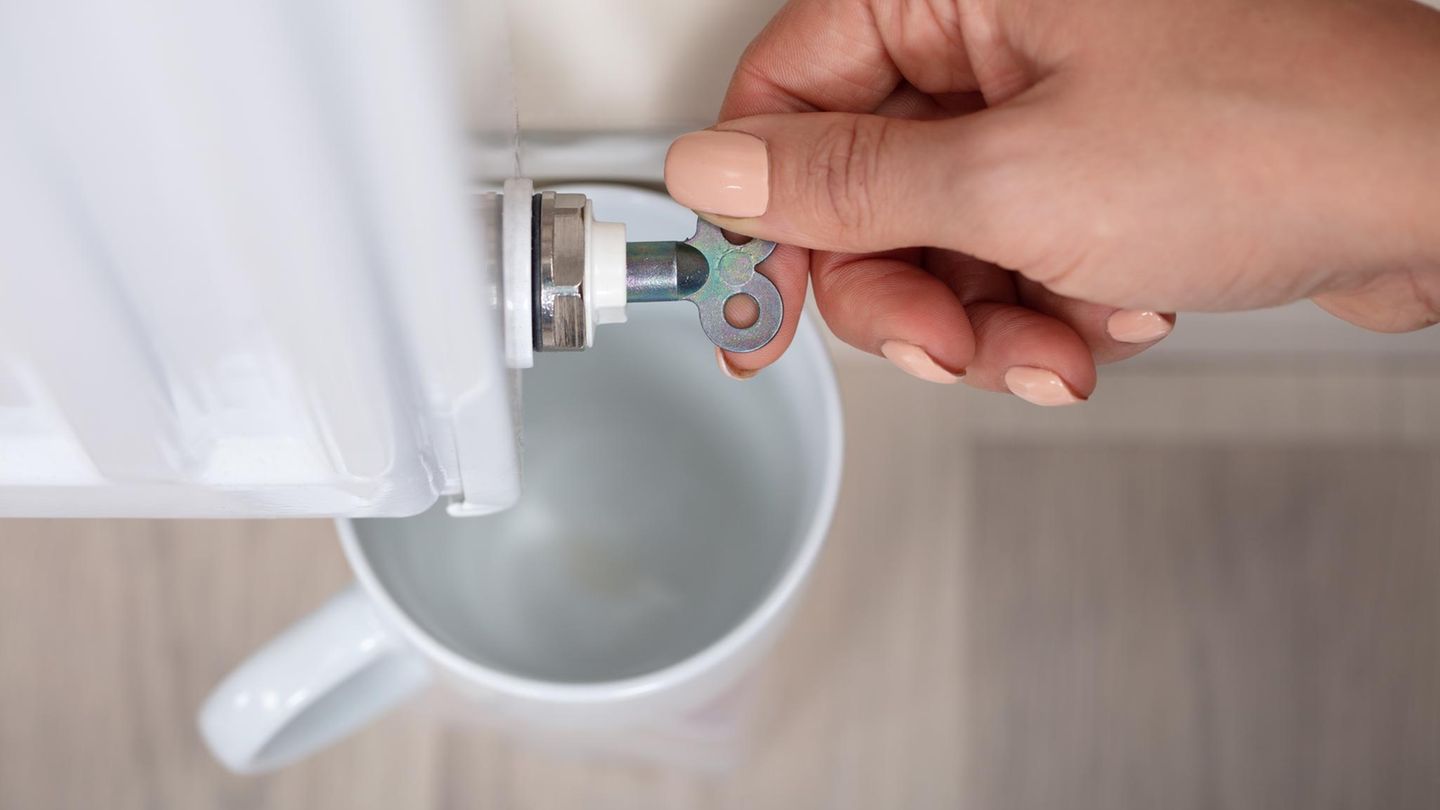 Nahaufnahme der Hand einer Person, die das Heizkörperentlüftungsventil dreht, um Luft mit Becher abzulassen