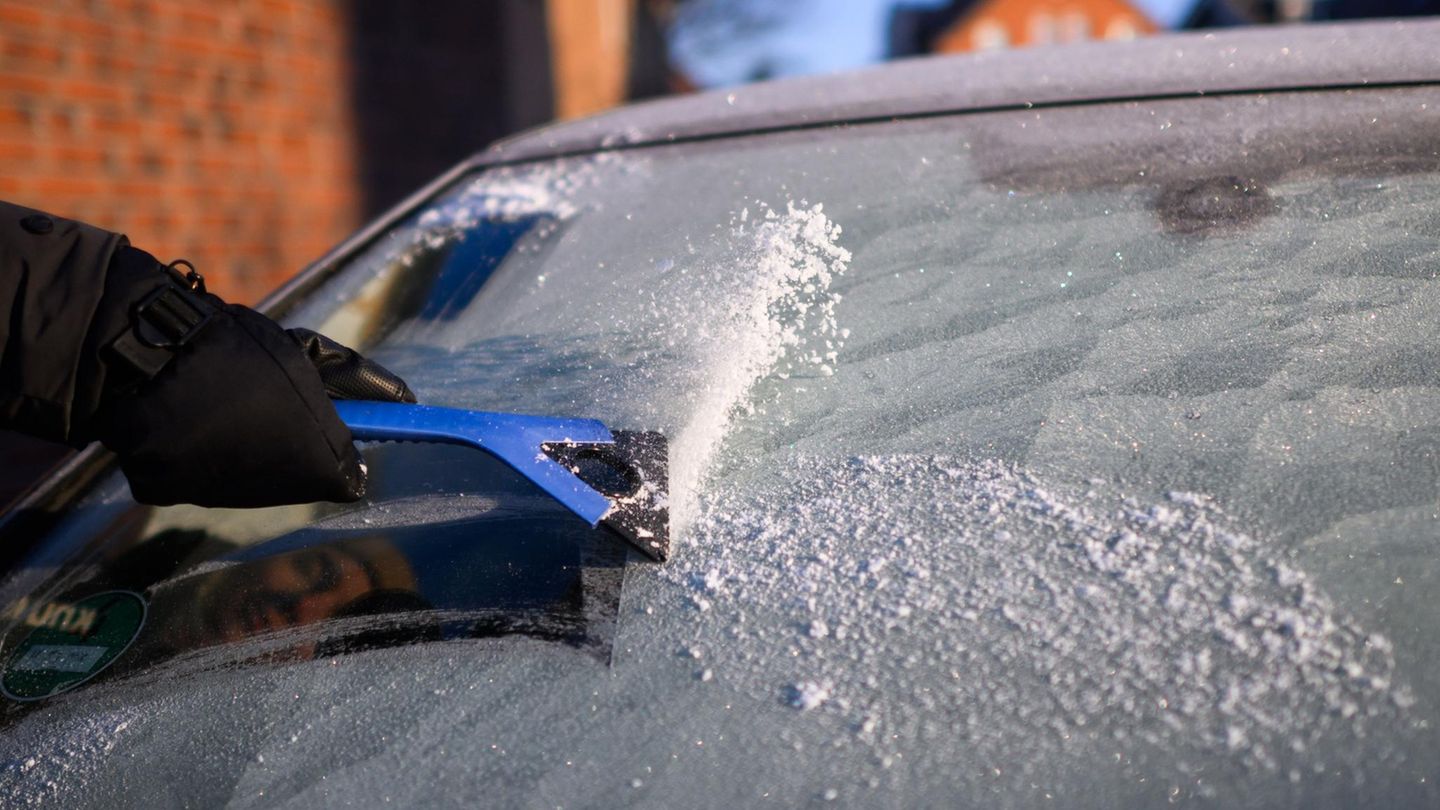 Eine vereiste Windschutzscheibe. Eine Hand im schwarzen Handschuh kratzt sie mit einem blauen Eiskratzer frei.
