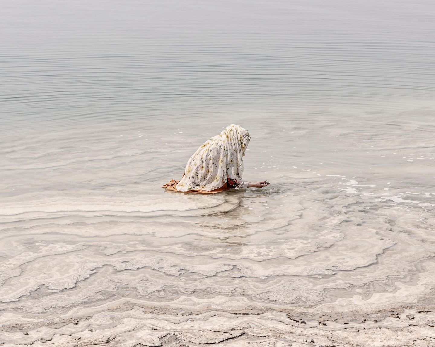 Am Ufer des Toten Meeres, in Jordanien, hockt eine Frau im flachen Wasser und tastet nach Salzsteinen. Beinahe entrückt erscheint die Szene und sie ist Teil einer Jahrtausende alten Tradition: Den Mineralen des hypersalinen Sees wird eine reinigende und heilende Wirkung zugeschrieben. Das Salz soll die Haut beruhigen, die Muskeln entspannen, kleine Wunden versiegeln. In Jordanien gehören Rituale der Pflege und Reinigung für viele zum Alltag, wobei sie heute auch in die Wellnessindustrie und den Tourismus reichen.   Die Aufnahme ist Teil des internationalen "Walk of Water"–Fotowettbewerbs von UNESCO und Onewater. Die ausgezeichneten Bildgeschichten zeigen, wie eng Wasser mit Identität und Kultur verwoben ist – von rituellen Gesten bis zum alltäglichen Überleben