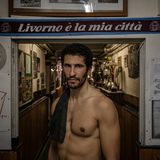 Im historischen Stadtteil Borgo Cappuccini im italienischen Livorno steht Karim Rachedi vor dem Eingang eines lokalen Clubs. Der Blick des Athleten wirkt ruhig. Und  dabei auch ein Stückweit herausfordernd. "Livorno ist meine Stadt" prangt auf dem Schal hinter dem Ruderer; ein kurzer Satz, der sportliche Disziplin mit Zugehörigkeit verknüpft. Denn das Rudern fordert hier nicht bloß Wettkampfsgeist. Es prägt auch Identität: ein kollektives Ritual zwischen Hafen und Meer, Wohnhäusern und Vereinsraum. Die Pokale im Hintergrund erzählen von Siegen, Jubel, Tradition