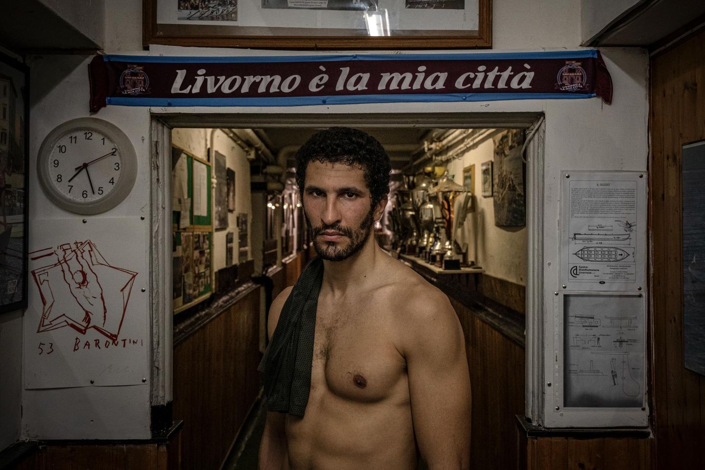 Im historischen Stadtteil Borgo Cappuccini im italienischen Livorno steht Karim Rachedi vor dem Eingang eines lokalen Clubs. Der Blick des Athleten wirkt ruhig. Und  dabei auch ein Stückweit herausfordernd. "Livorno ist meine Stadt" prangt auf dem Schal hinter dem Ruderer; ein kurzer Satz, der sportliche Disziplin mit Zugehörigkeit verknüpft. Denn das Rudern fordert hier nicht bloß Wettkampfsgeist. Es prägt auch Identität: ein kollektives Ritual zwischen Hafen und Meer, Wohnhäusern und Vereinsraum. Die Pokale im Hintergrund erzählen von Siegen, Jubel, Tradition