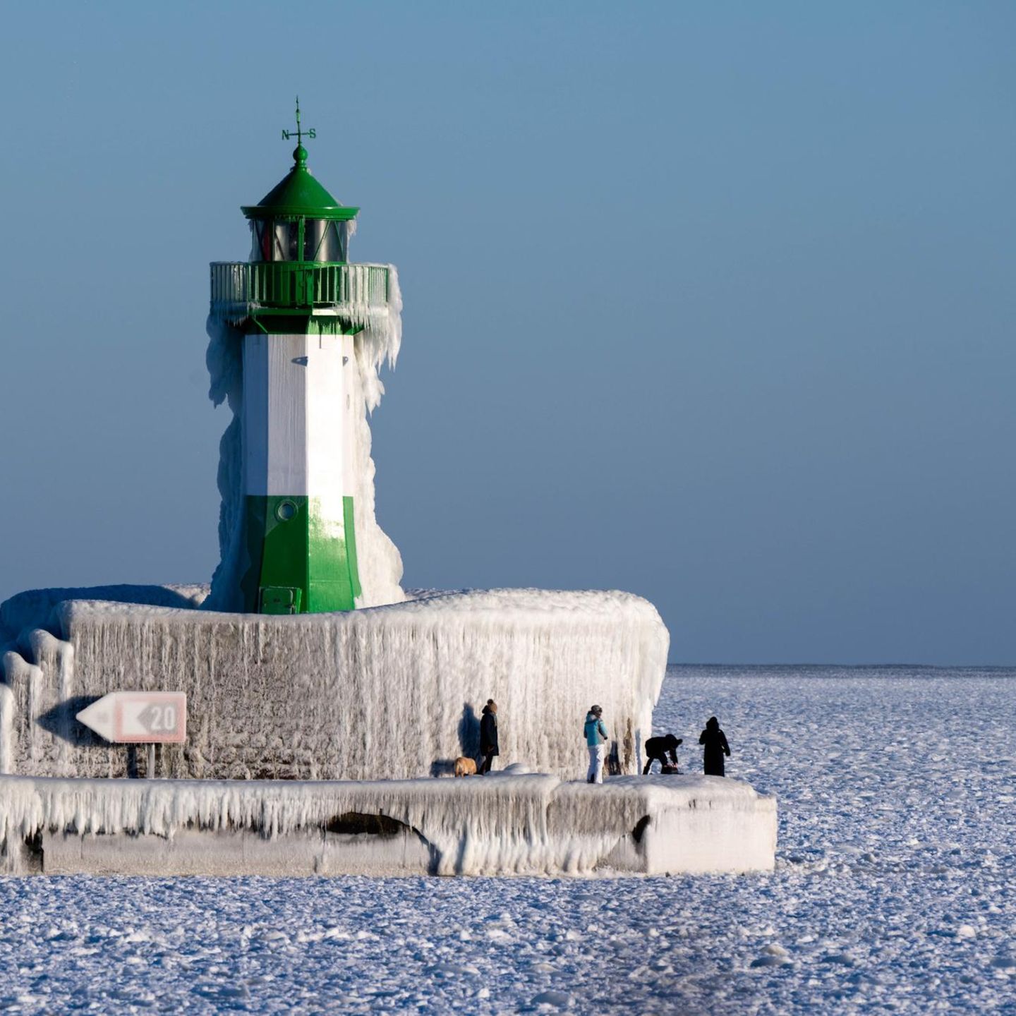 Grün-weißer Leuchtturm in der Ostsee. Lange Eiszapfen bedecken ihn. Die Ostsee ist bis zum Horizont gefroren.