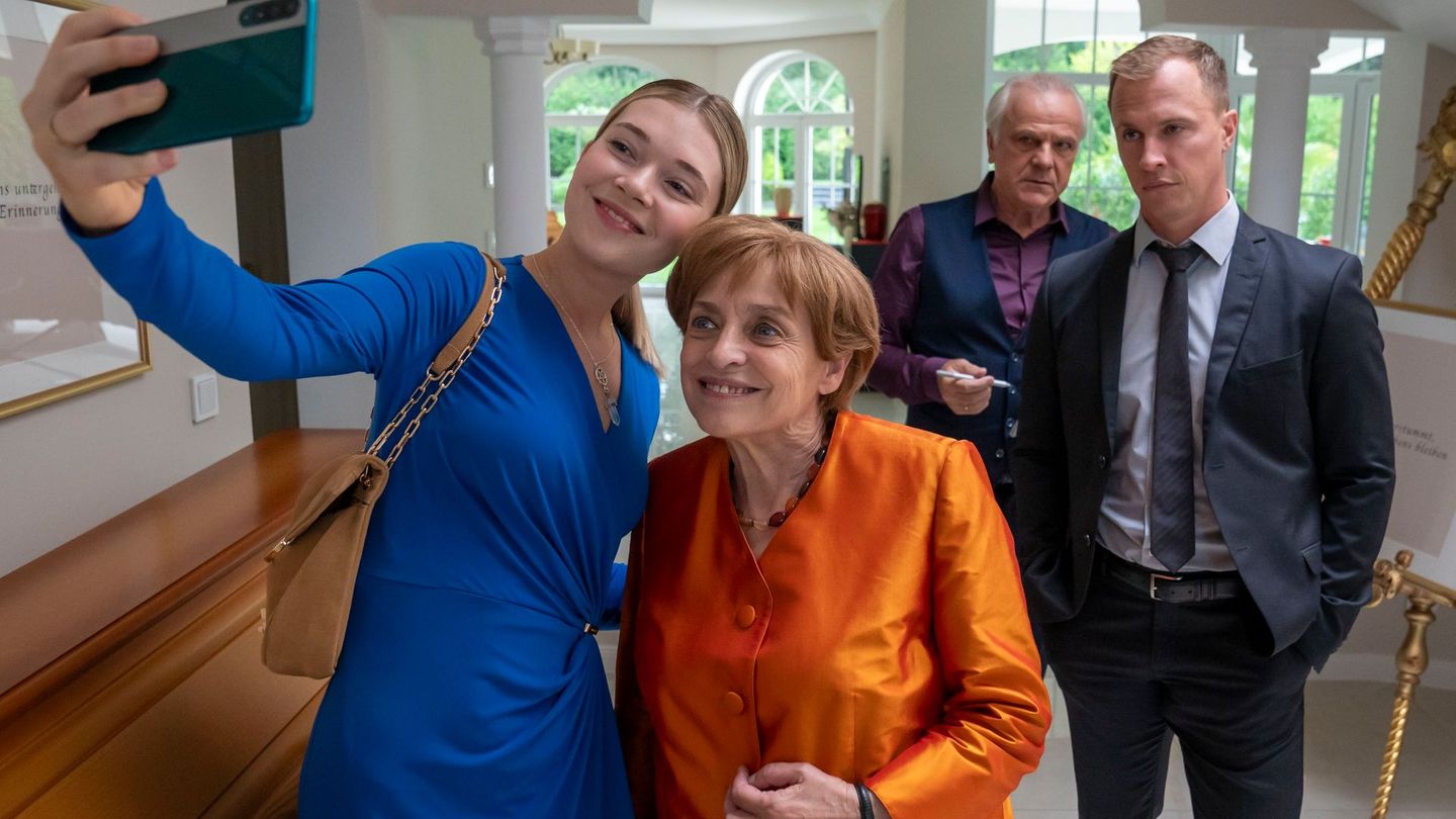 Charu Borscht (Farina Flebbe, links), die junge Frau des zweiten Bestatters im Ort, macht ein Selfie mit Angela Merkel (Katharina Thalbach). Ehemann und Bestatter Borscht (Bernhard Schütz) sowie Merkels Personenschützer Mike (Tim Kalkhof, rechts) schauen zu.