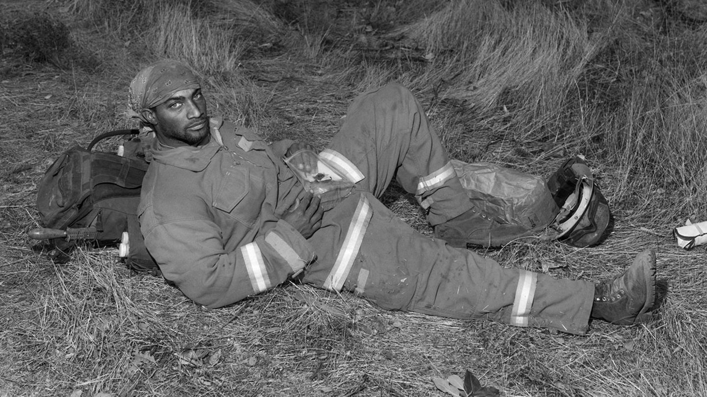 Ein Feuerwehrmann liegt im Gras auf seinem Rucksack, er schaut direkt in die Kamera
