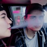 Junger Mann sitzt zusammen mit einer jungen Frau im Auto, sie raucht, er schaut in die Kamera.