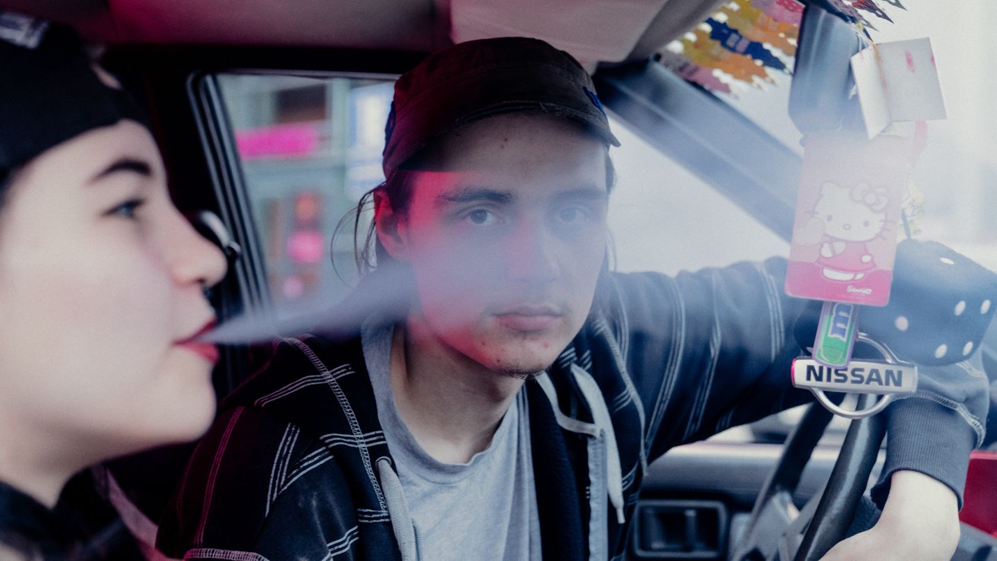 Junger Mann sitzt zusammen mit einer jungen Frau im Auto, sie raucht, er schaut in die Kamera.