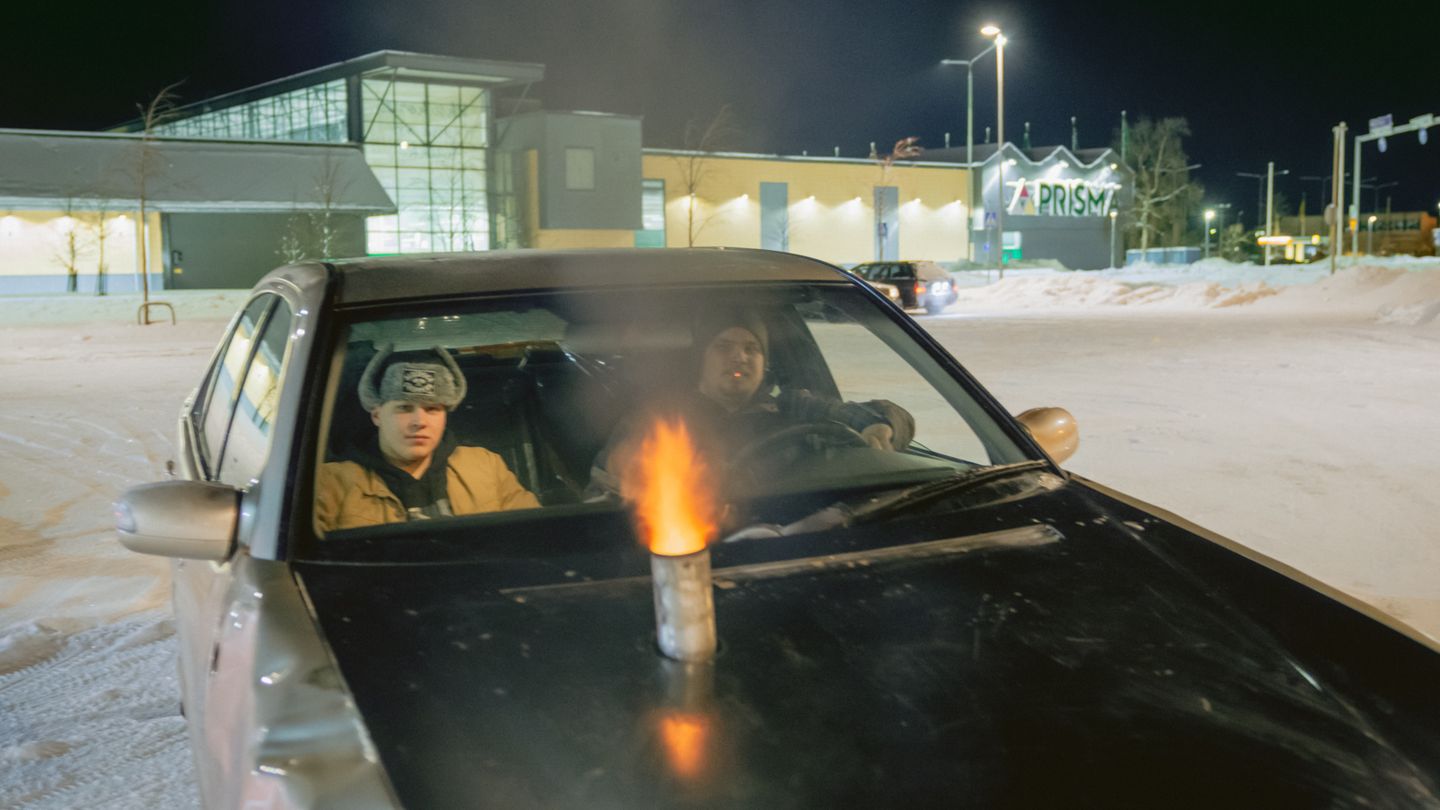 Zwei finnische Jugendliche in einem Auto mit einem Auspuff auf der Motorhaube, aus dem Flammen schlagen