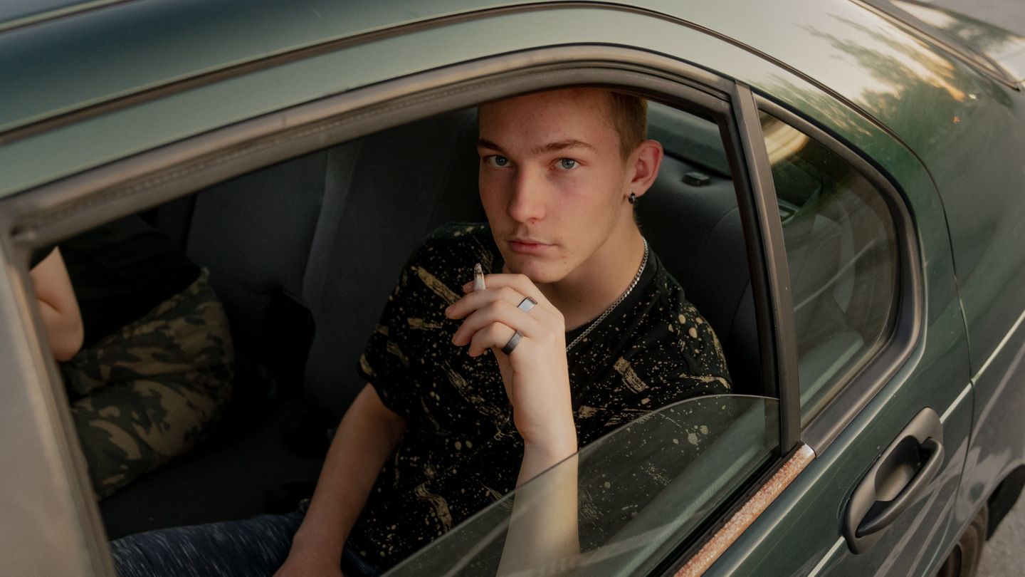 Ein junger Mann sitzt rauchend auf dem Rücksitz eines dunkelgrünen, rostigen Autos und schaut aus dem Fenster