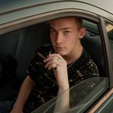 Ein junger Mann sitzt rauchend auf dem Rücksitz eines dunkelgrünen, rostigen Autos und schaut aus dem Fenster