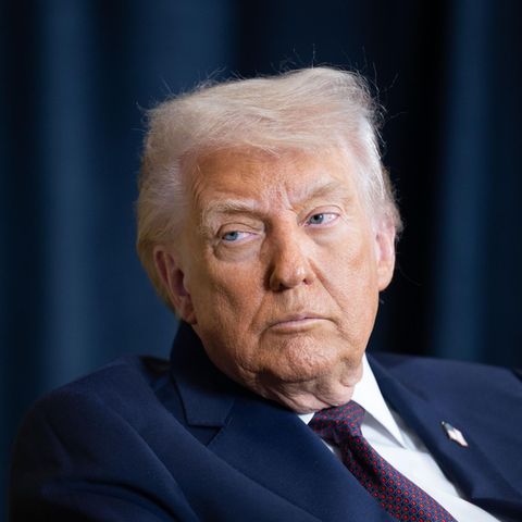 Donald Trump sitzt in einem blauen Anzug vor einem Blauen Vorhang und schaut unzufrieden