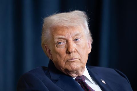 Donald Trump sitzt in einem blauen Anzug vor einem Blauen Vorhang und schaut unzufrieden