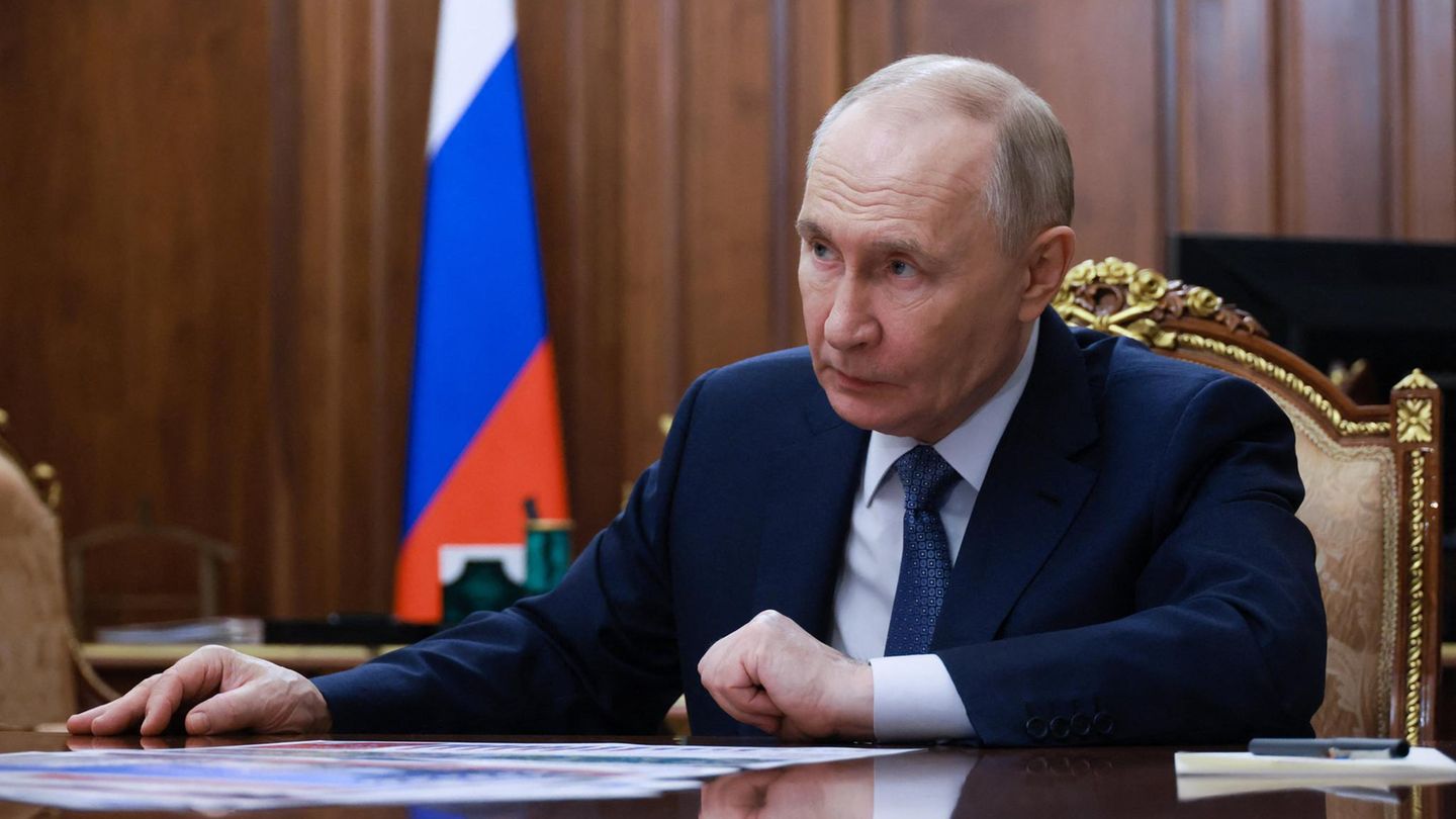 Putin sitzt an einem Tisch, die russische Flagge im Hintergrund