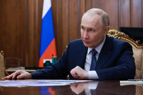 Putin sitzt an einem Tisch, die russische Flagge im Hintergrund