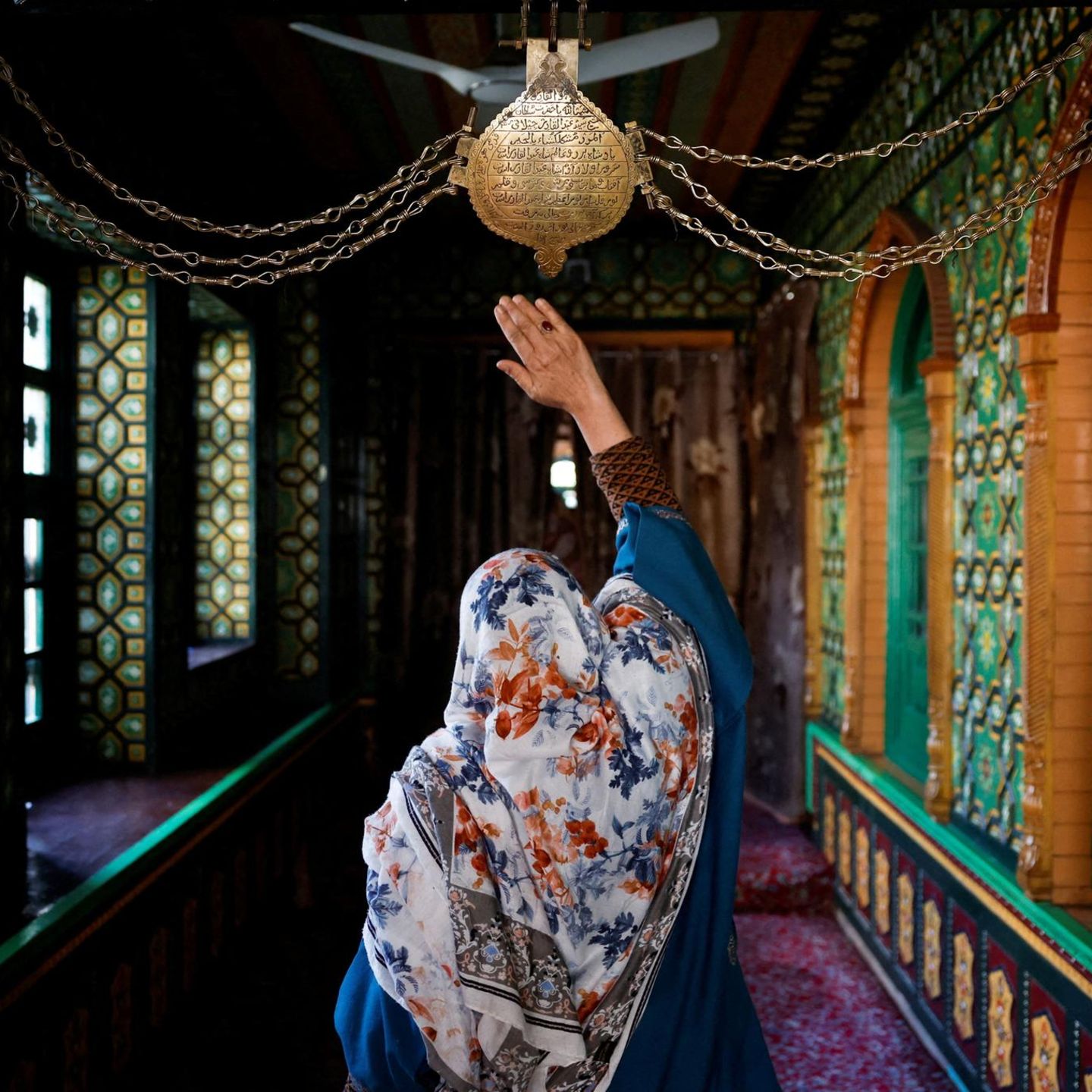 Srinagar, Indien. Diese gläubige Frau berührt ehrfürchtig die Kette am Schrein von Sheikh Abdul Qadir Jeelani. Umgeben von leuchtenden Mustern und Farben sucht sie während des Ramadan nach Spiritualität. 