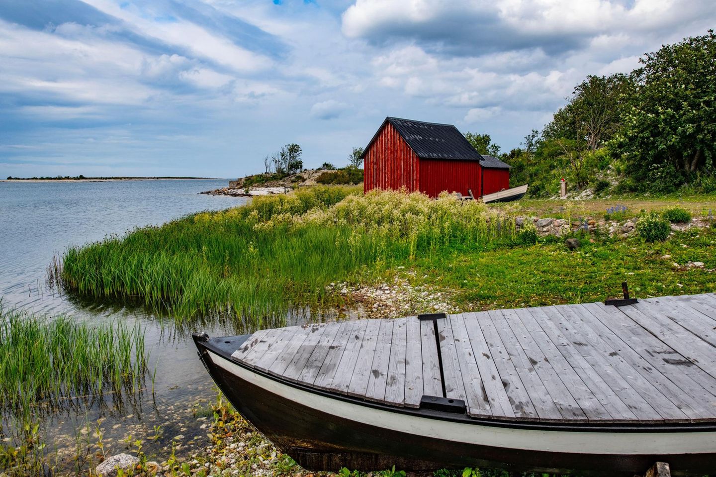 Im Vordergrund ein Holzboot, im Hintergrund ein rotes Holzhaus an einem See gelegen