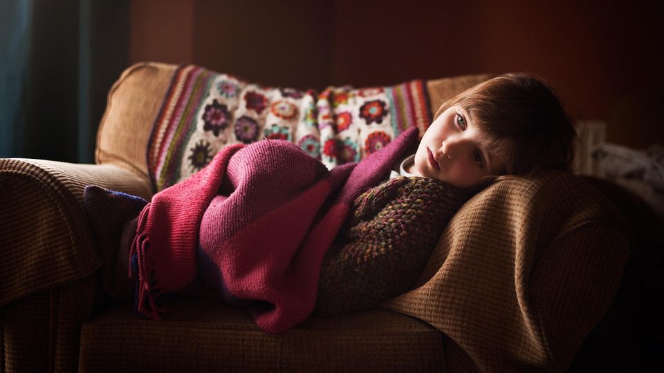 Ein junges Kind liegt eingekuschelt auf einem Sessel und schaut traurig