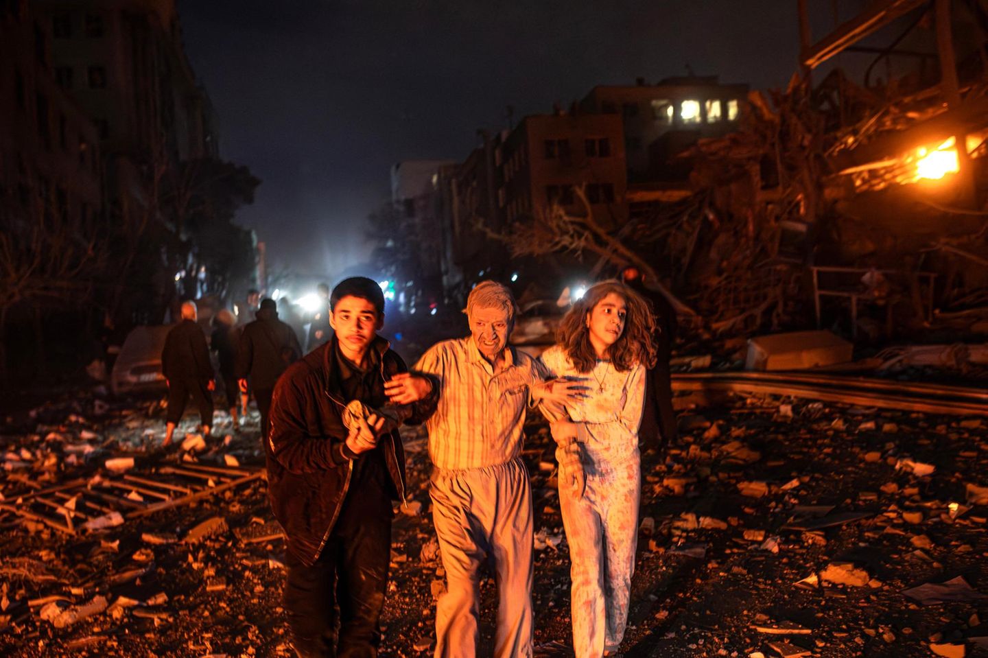 Drei junge Leute laufen bei Nacht in Pyjamas auf einer zerstörten Straße. Sie sind staubbedeckt.