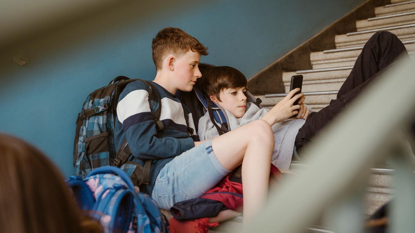 zwei Jungen in der Pubertät flezen auf einer Steintreppe in einem Gebäude und schauen auf ein Smartphone