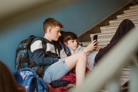 zwei Jungen in der Pubertät flezen auf einer Steintreppe in einem Gebäude und schauen auf ein Smartphone