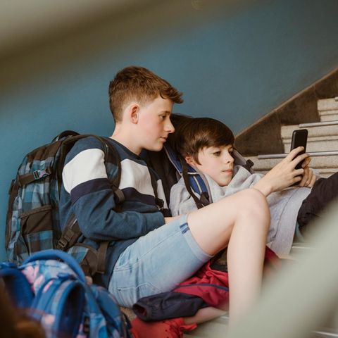 zwei Jungen in der Pubertät flezen auf einer Steintreppe in einem Gebäude und schauen auf ein Smartphone