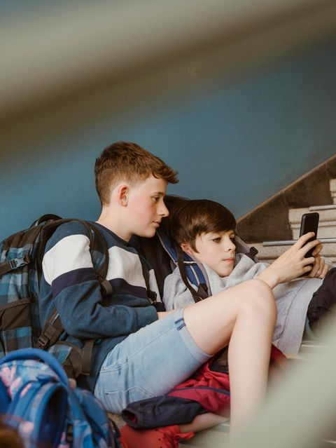zwei Jungen in der Pubertät flezen auf einer Steintreppe in einem Gebäude und schauen auf ein Smartphone