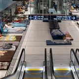 Matratzen, Kissen und Decken liegen an den Rändern der Bahnstation. Von oben ist die Rolltreppe und die Halle zu sehen.