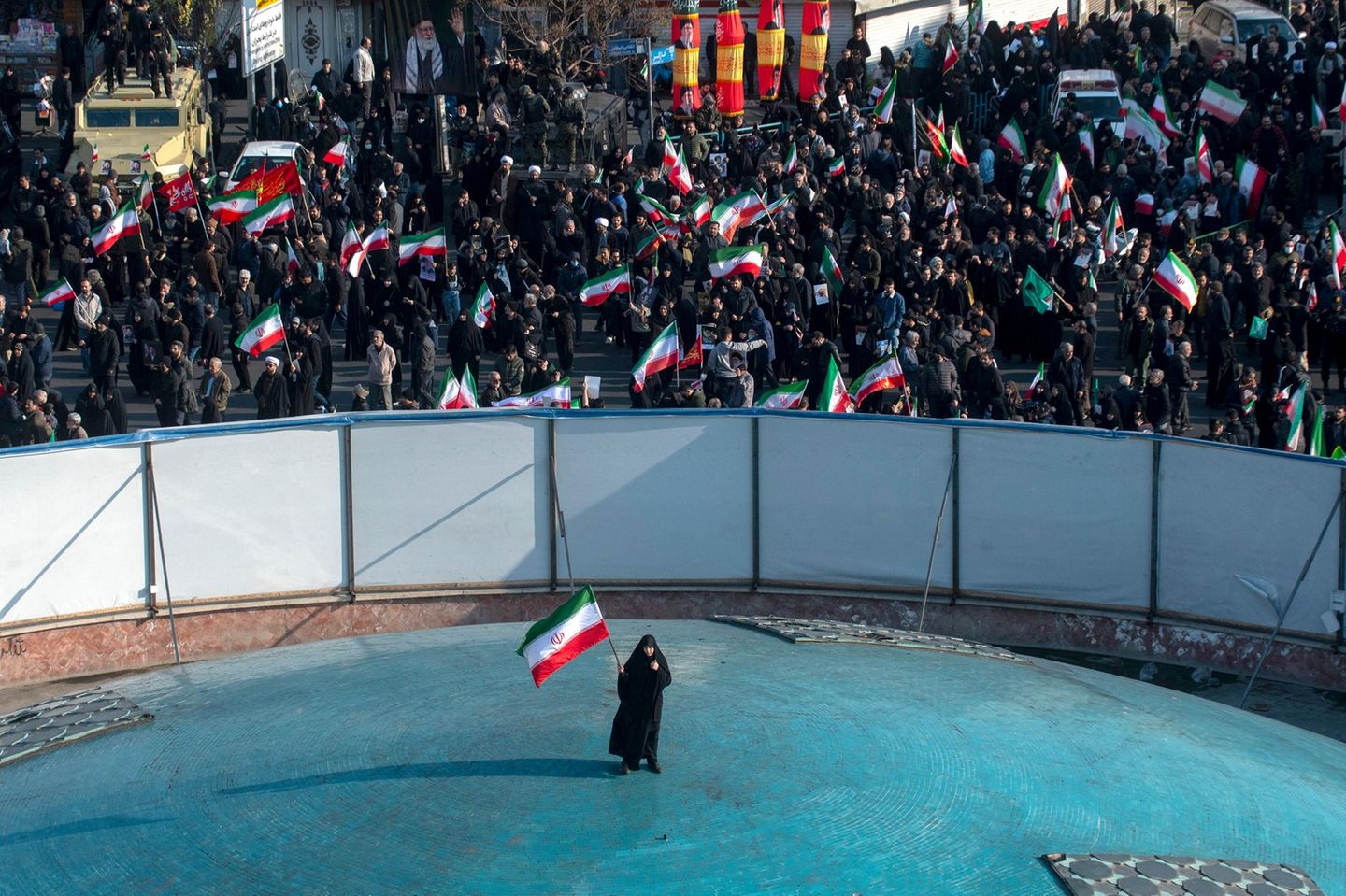 Iranische Frau steht alleine in der Mitte mit einer Iranflagge und demonstriert. Um sie herum eine Absperrung mit weiteren.
