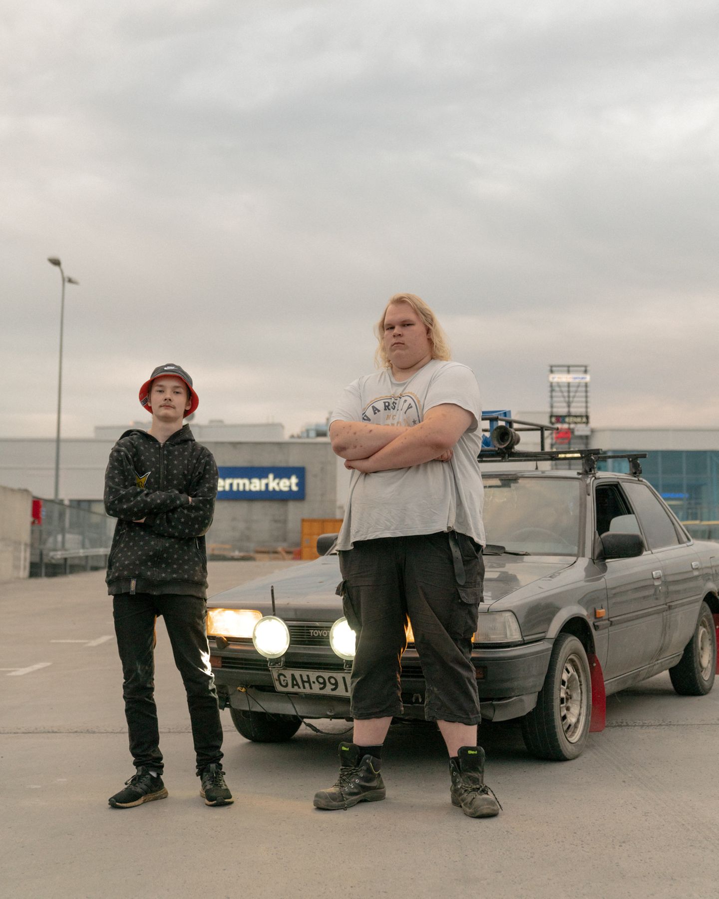 Zwei Jungs stehen mit verschrenkten Armen vor einem grauen Auto (Toyota)