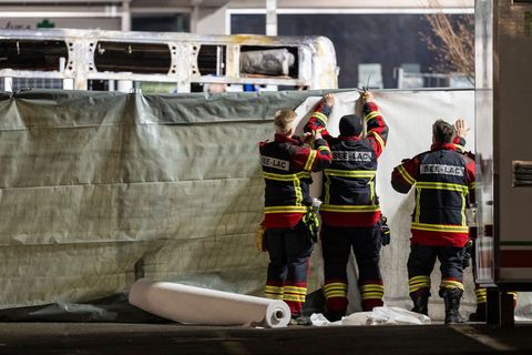 Feuerwehrleute verdecken den ausgebrannten Bus. Foto: Alessandro Della Valle/KEYSTONE/dpa