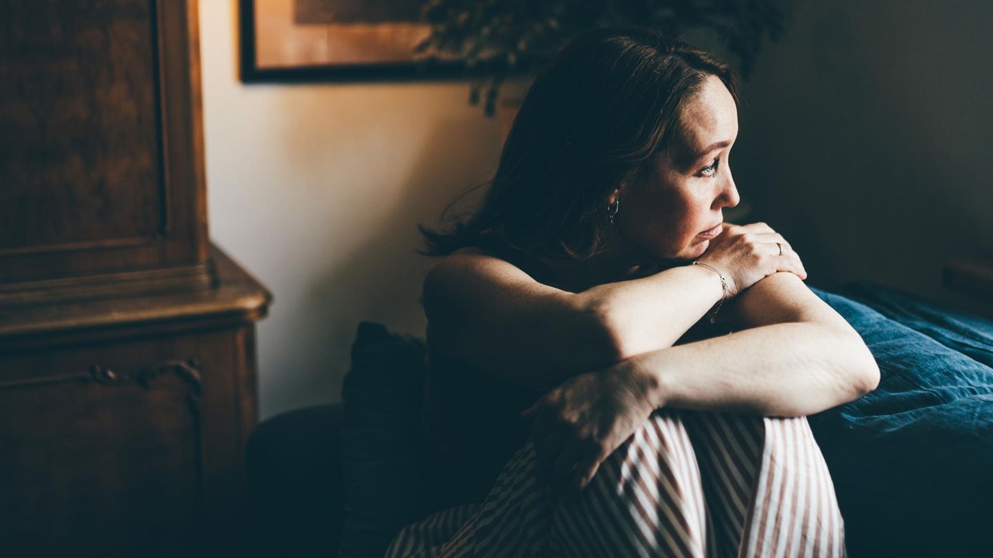 Symbolfoto Affäre: Eine Frau sitzt mit angezogenen Knien in einem Zimmer und blickt traurig zur Seite