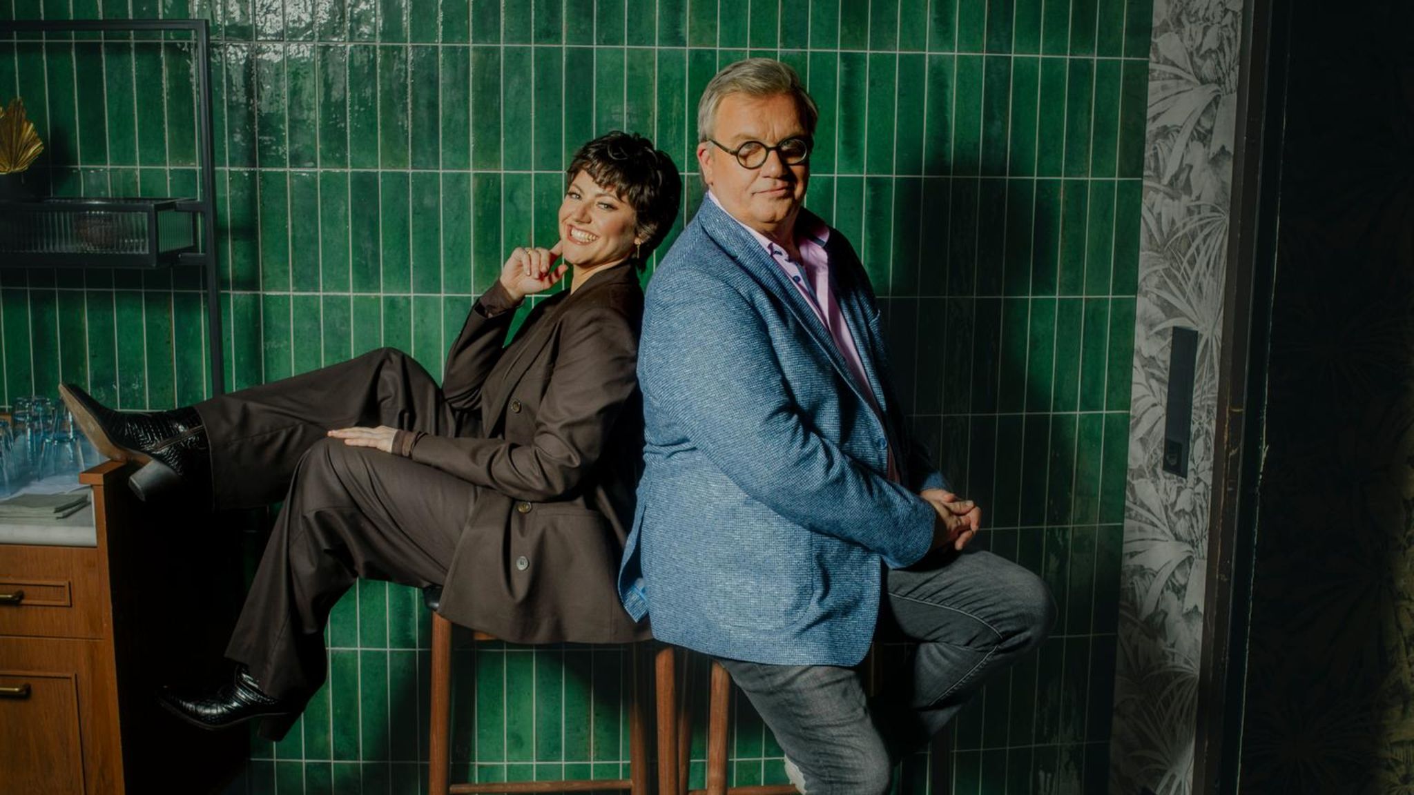 Hape Kerkeling und Tahnee sitzen Rücken an Rücken auf Barhockern vor einer grün gefließten Wand