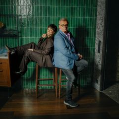 Hape Kerkeling und Tahnee sitzen Rücken an Rücken auf Barhockern vor einer grün gefließten Wand