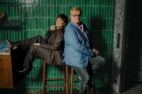 Hape Kerkeling und Tahnee sitzen Rücken an Rücken auf Barhockern vor einer grün gefließten Wand