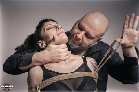 Eine schwarzhaarige Frau lehnt an einem Mann mit Bart, der ihren Oberkörper fesselt. Sie praktizieren Shibari.